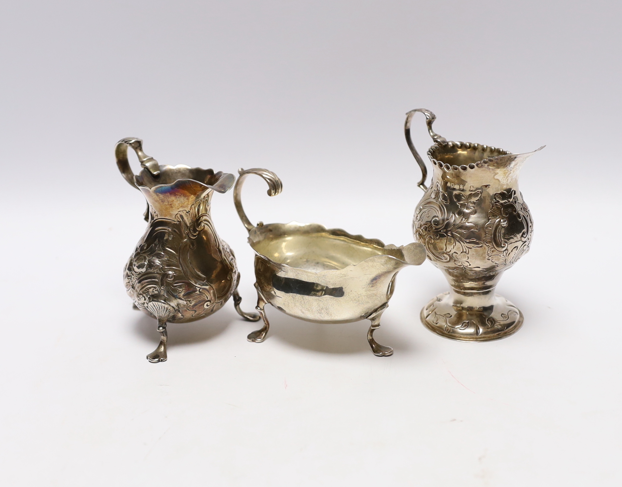 A late George II silver sauceboat with flying scroll handled, London, 1759 and two Georgian silver cream jugs, with later embossed decoration (a.f.)